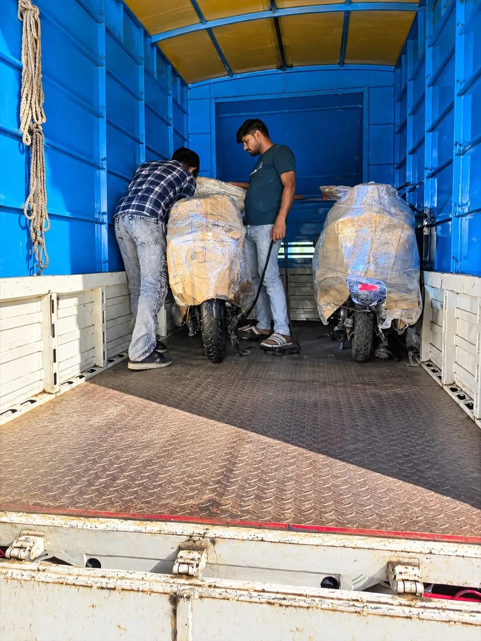 Two workers secure motorcycles wrapped in protective padding inside a blue truck